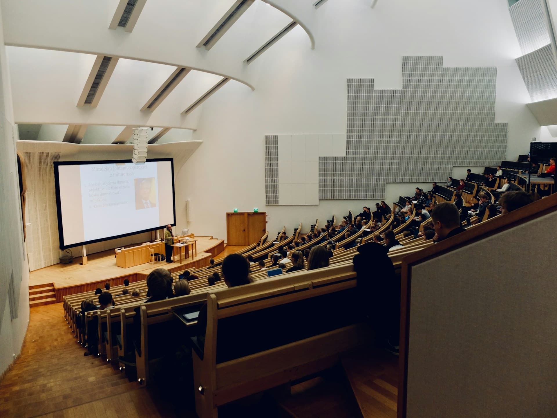 階段状の座席が並ぶ大学の大講義室で、学生たちがスクリーンを使った講義を受けている様子