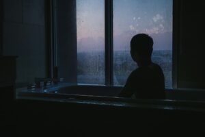 夕暮れ時、湯船に浸かりながら曇った窓の外を眺める人物のシルエット