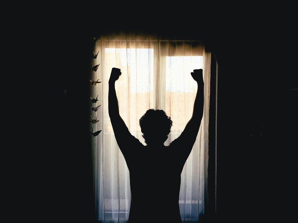 朝の窓辺で両腕を伸ばす人のシルエット。朝は起きただけで偉いと考えるイメージ